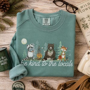 May include: A sage green crewneck sweatshirt featuring a graphic of a raccoon, a bear, and a fox in a forest setting with a campfire and the text "be kind to the locals" printed in white script. The garment has a Comfort Colors brand label.