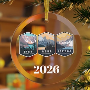 May include: A clear, round Christmas ornament with a gold ribbon. The ornament features three rectangular images of Banff, Jasper, and Kootenay National Parks. The year "2026" is printed in white below the images.