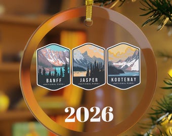 Puede incluir: Un adorno navideño redondo y transparente con una cinta dorada. El adorno presenta tres imágenes rectangulares de los parques nacionales de Banff, Jasper y Kootenay. El año "2026" está impreso en blanco debajo.