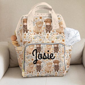 May include: A beige and cream diaper bag with a floral and Highland cow pattern. The bag has a top handle, adjustable straps, and multiple pockets. The name "Josie" is written in black script on the front. A wooden teething ring and a white blanket are attached.