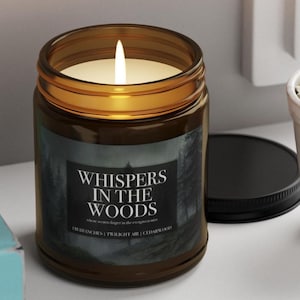 May include: A lit candle in a brown glass jar with a black lid. The label reads "WHISPERS IN THE WOODS" with additional text. The candle is on a white surface.