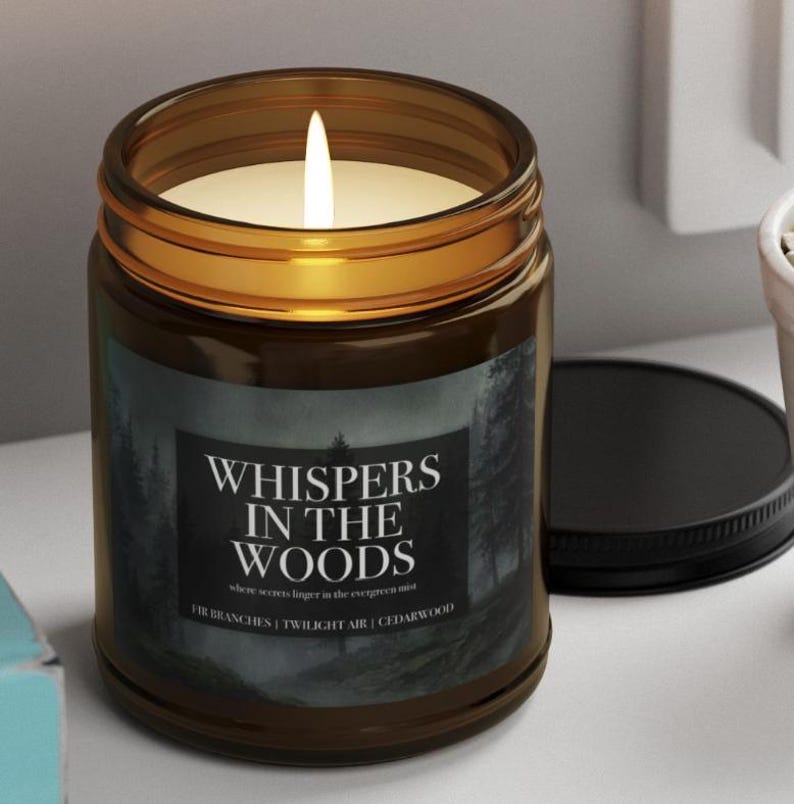 May include: A lit candle in a brown glass jar with a black lid. The label reads "WHISPERS IN THE WOODS" with additional text. The candle is on a white surface.