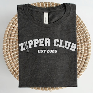 May include: A dark gray t-shirt with the text "ZIPPER CLUB EST 2026" in white, featuring a zipper graphic. The shirt is folded and placed on a round, woven basket.