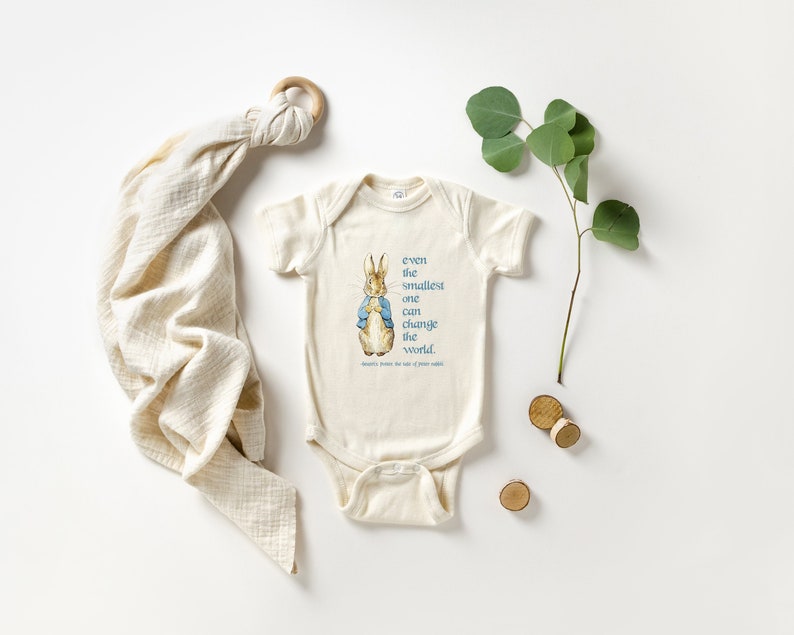 May include: A cream-colored baby onesie featuring a Peter Rabbit illustration and the quote, "even the smallest one can change the world." A muslin swaddle blanket and eucalyptus sprig are also present.