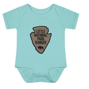 May include: A light turquoise short-sleeved baby bodysuit featuring a tan arrowhead-shaped graphic on the chest. The graphic contains the black text "LITTLE NATIONAL PARK RANGER" and a small black silhouette of a bison at the bottom centre.
