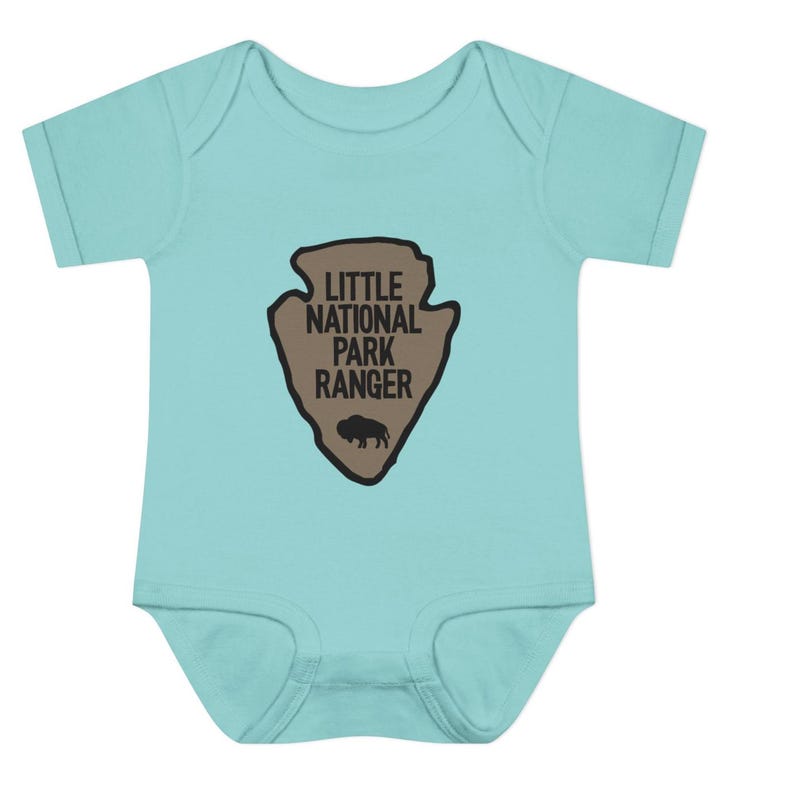 May include: A light turquoise short-sleeved baby bodysuit featuring a tan arrowhead-shaped graphic on the chest. The graphic contains the black text "LITTLE NATIONAL PARK RANGER" and a small black silhouette of a bison at the bottom centre.