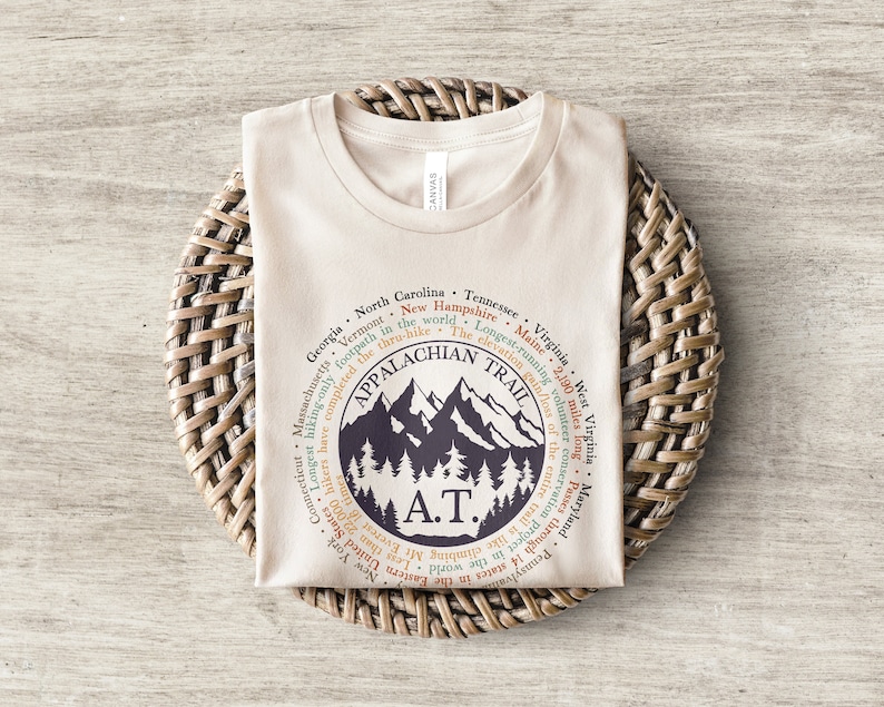 Op de afbeelding: Een beige T-shirt met een grafisch ontwerp van de Appalachian Trail. Het ontwerp bevat de tekst "Appalachian Trail" en "A.T." samen met een bergketen en bomen. De tekst rond het ontwerp geeft de staten weer die de route doorkruist.