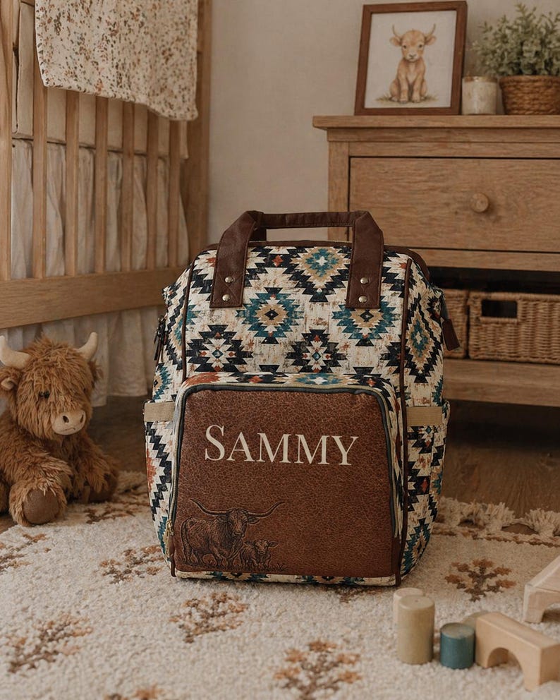 May include: A diaper bag with a brown leather front panel displaying the name "SAMMY" and a bull illustration. The bag features a geometric pattern in shades of blue, white, and brown. A stuffed animal and framed artwork are visible in the background.
