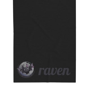 May include: Black blanket featuring a cartoon bat illustration in a circle with a moon and stars. The word "raven" is printed in a dark gray font. The blanket is a solid black color.