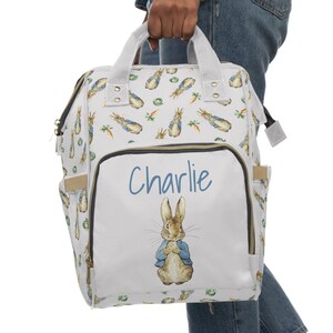 May include: A white diaper bag with a Peter Rabbit design. The bag features a front pocket with the name "Charlie" in blue script and a watercolor illustration of Peter Rabbit. The bag has multiple pockets and a top handle.