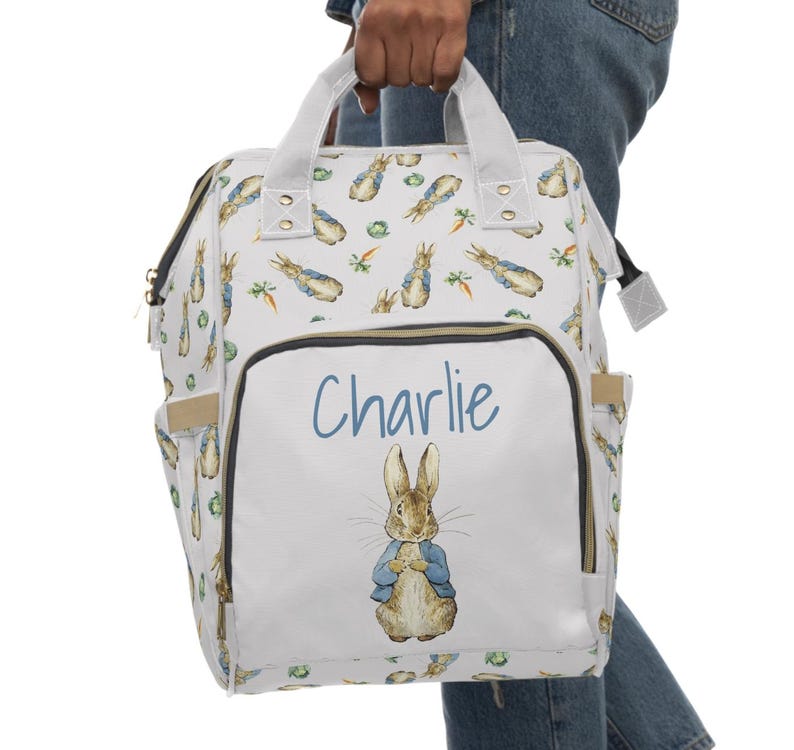 May include: A white diaper bag with a Peter Rabbit design. The bag features a front pocket with the name "Charlie" in blue script and a watercolor illustration of Peter Rabbit. The bag has multiple pockets and a top handle.