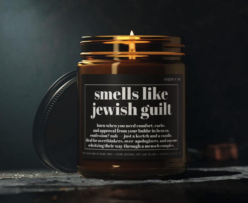 May include: A lit candle in an amber glass jar with a black label. The label features white text reading: smells like jewish guilt, burn when you need comfort, carbs, and approval from your bubbe in heaven. confession? nah&mdash;just a kvetch and a candle.