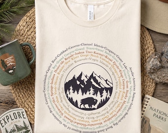 May include: A cream-colored t-shirt featuring a circular graphic design. The center shows a dark silhouette of mountains, pine trees, and a bison. Surrounding the central image are the names of various United States National Parks in text.