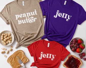 May include: Three t-shirts in tan, purple, and red, each with a different white graphic. The tan shirt says "peanut butter", the purple shirt says "jelly", and the red shirt says "jelly". The image also includes a bowl of peanuts, a slice of bread with peanut butter, and a slice of bread with jelly.