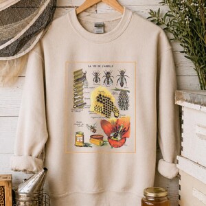 May include: Cream-coloured sweatshirt with a vintage bee and honeycomb illustration, and the text "LA VIE DE L'ABEILLE". The graphic includes detailed drawings of bees, honey, and a red flower. A jar of raw honey is also visible.