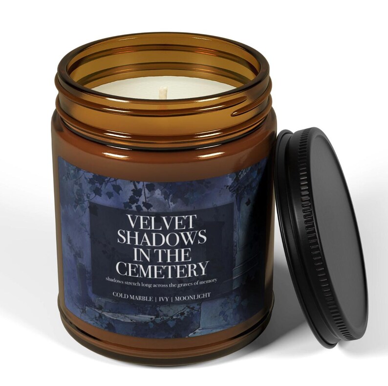 May include: A brown glass jar candle with a black lid. The candle has a white wax interior and a label that reads "VELVET SHADOWS IN THE CEMETERY". The label also includes the phrases "COLD MARBLE | IVY | MOONLIGHT".