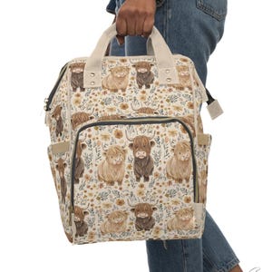 May include: A cream-colored backpack featuring a repeating pattern of Highland cows in various shades of brown. The bag has a floral design, a front pocket, side pockets, and a top handle. The straps and handle are a light beige color.