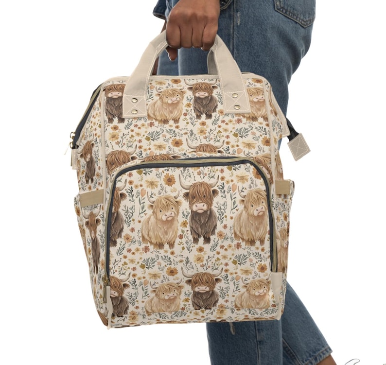 May include: A cream-colored backpack featuring a repeating pattern of Highland cows in various shades of brown. The bag has a floral design, a front pocket, side pockets, and a top handle. The straps and handle are a light beige color.