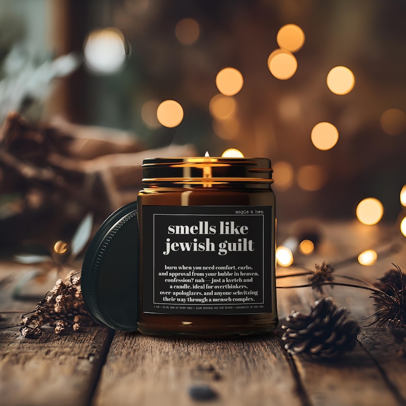 May include: A brown glass jar candle with a black lid and a label that reads: smells like jewish guilt. Burn when you need comfort, carbs, and approval from your bubbe in heaven, confession? nah &mdash; just a kvetch and a candle.