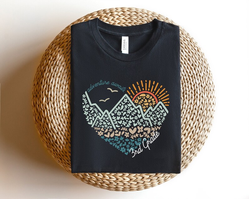 May include: A black t-shirt featuring a heart-shaped mountain design with the text "adventure awaits" and "3rd Grade." The design includes mountains, flowers, birds, and a sun in shades of orange, teal, and beige.