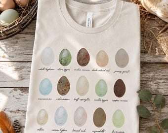 May include: A cream-colored t-shirt featuring a graphic print of fifteen distinct chicken eggs arranged in three rows. Each egg is labeled with its breed name, including White Leghorn, Olive Egger, Cuckoo Maran, Rhode Island Red, Jersey Giant, Ameraucana, Welsummer, Buff Orpington, Easter Egger, Copper Maran, Silkie, Cream Legbar, Barred Rock, Wyandotte, and Favaucana.