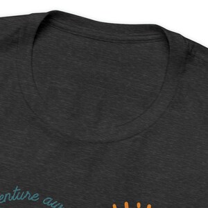 May include: Dark gray t-shirt with a crew neck. The shirt features the text "venture awaits" in teal script, with an orange sun graphic. The fabric has a heathered texture.