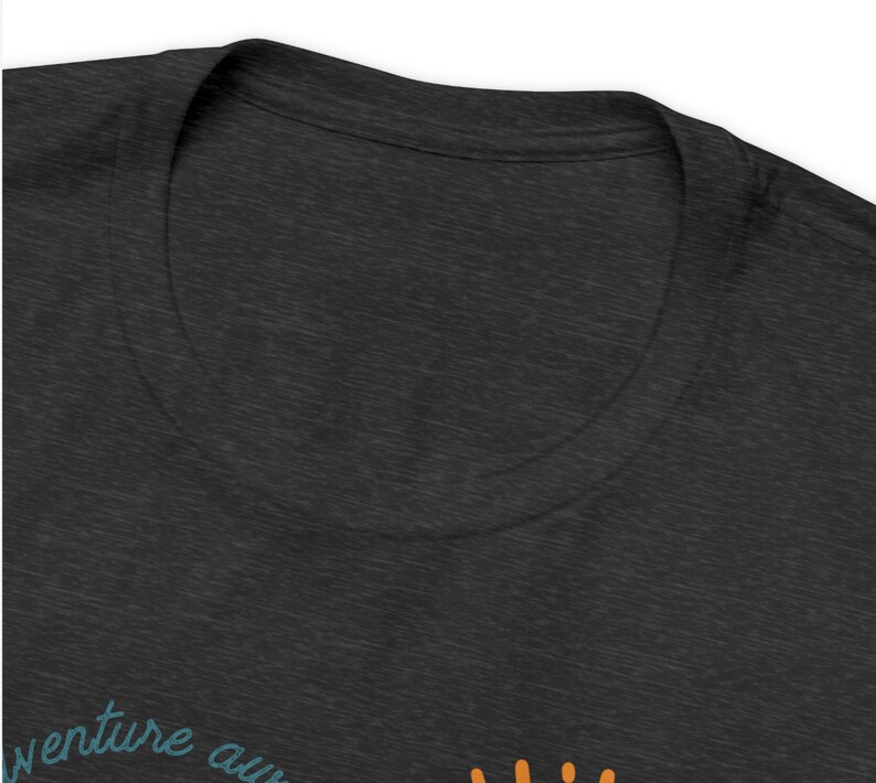 May include: Dark gray t-shirt with a crew neck. The shirt features the text "venture awaits" in teal script, with an orange sun graphic. The fabric has a heathered texture.