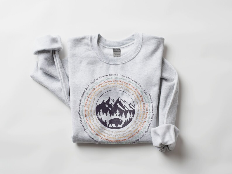 May include: A light grey crewneck sweatshirt featuring a circular graphic on the chest. The design includes a dark purple silhouette of mountains, pine trees, and a bison, surrounded by a ring of text listing various national park names.
