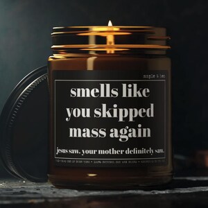 May include: A lit candle in a brown glass jar with a black lid. The label reads "smells like you skipped mass again" with the text "jesus saw, your mother definitely saw." The jar is labeled "maple & ben".