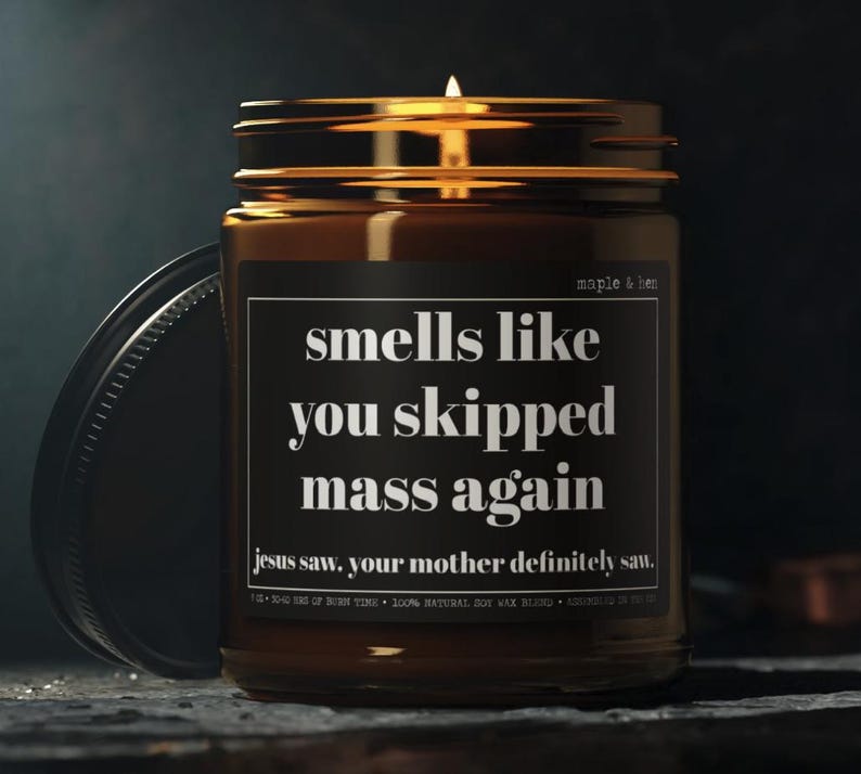 May include: A lit candle in a brown glass jar with a black lid. The label reads "smells like you skipped mass again" with the text "jesus saw, your mother definitely saw." The jar is labeled "maple & ben".