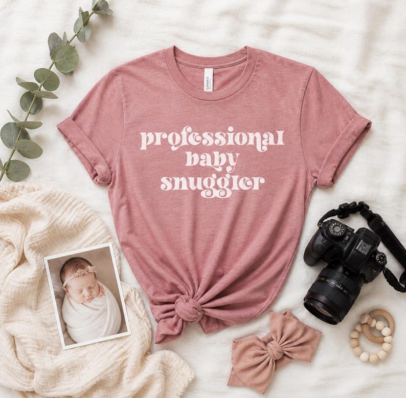 May include: A dusty rose t-shirt with the text "professional baby snuggler" in white script. The shirt is knotted at the waist. A baby photo, camera, bow, and wooden teething ring are also in the image.