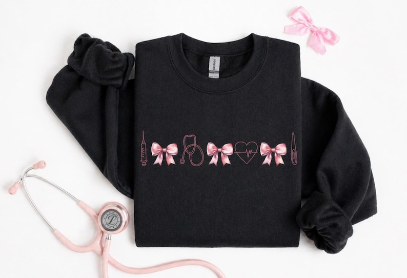 May include: Black sweatshirt with pink medical-themed embroidery. The design includes a syringe, bows, a stethoscope, a heart with a heartbeat line, and a thermometer. A pink bow and a stethoscope are also visible.