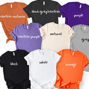 May include: A collection of eight short-sleeve crew neck t-shirts in various colors including heather autumn, dark grey heather, purple, heather purple, natural, grey, black, white, and orange, each labeled with its respective color name in white text.