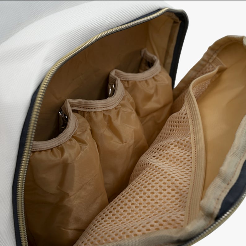 May include: Open view of a white and black travel bag interior with multiple compartments. The bag features a gold zipper, three tan fabric pockets, and a mesh pocket. The interior is tan, and the bag's exterior is white and black.