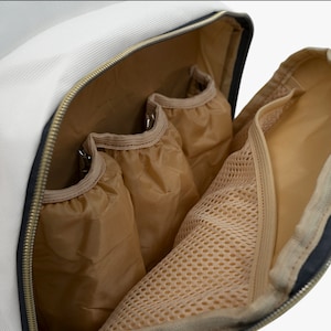 May include: Open view of a white and black travel bag interior with multiple compartments. The bag features a gold zipper, three tan fabric pockets, and a mesh pocket. The interior is tan, and the bag's exterior is white and black.