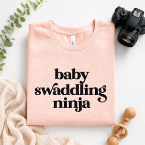 Puede incluir: Una camiseta rosa claro con cuello redondo con las palabras "baby swaddling ninja" impresas en negro. La camiseta se muestra sobre una superficie blanca con una c&aacute;mara, un sonajero de madera y vegetaci&oacute;n en el fondo.