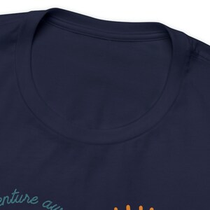 May include: Navy blue t-shirt with the words "adventure awaits" in a teal script. The shirt has a crew neck and is made of a soft material. The text is partially visible at the bottom of the image.