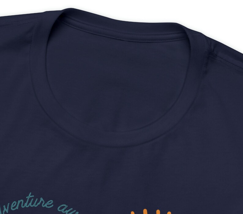 May include: Navy blue t-shirt with the words "adventure awaits" in a teal script. The shirt has a crew neck and is made of a soft material. The text is partially visible at the bottom of the image.