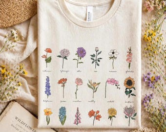 May include: A cream-colored crew neck t-shirt featuring a botanical print of eighteen distinct flowers arranged in three rows. Each flower is labeled with its name in cursive script, including anthurium, hydrangea, aster, rose, anemone, gladiolus, delphinium, freesia, carnation, cowslip, orchid, sunflower, and others.