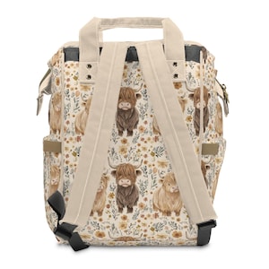 May include: A cream-colored backpack with a repeating pattern of Highland cows and floral elements in shades of brown, tan, and yellow. The backpack has beige straps and a top handle, with black accents on the bottom and sides.