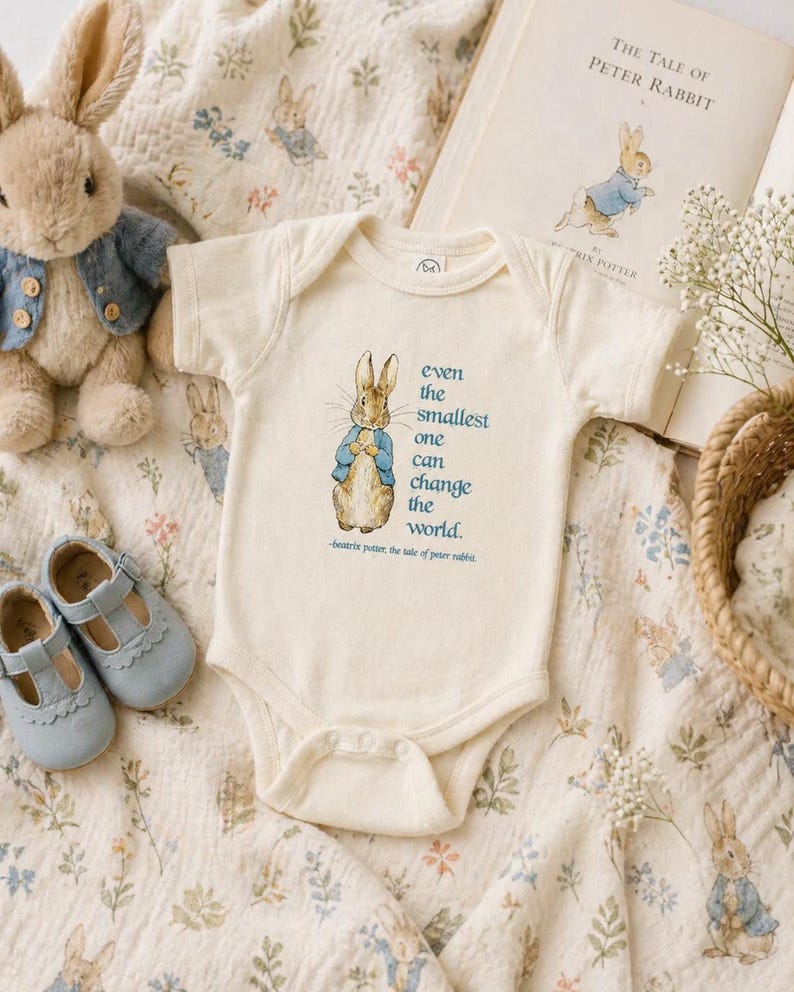 May include: A cream-colored baby onesie featuring a Peter Rabbit illustration and the quote, "even the smallest one can change the world." The onesie is surrounded by a plush rabbit, baby shoes, and a book titled "The Tale of Peter Rabbit."