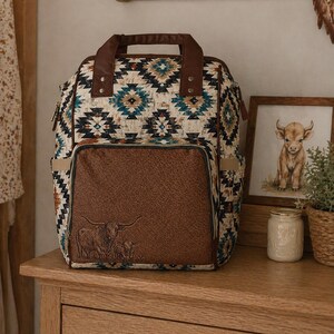 May include: A patterned backpack with a brown leather front pocket featuring an embossed image of a cow and calf. The bag has a brown handle and straps, and a geometric pattern in shades of blue, beige, and brown. It sits on a wooden surface.