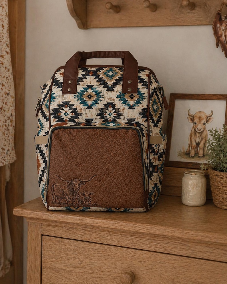May include: A patterned backpack with a brown leather front pocket featuring an embossed image of a cow and calf. The bag has a brown handle and straps, and a geometric pattern in shades of blue, beige, and brown. It sits on a wooden surface.