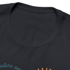 May include: Dark navy blue t-shirt with the words "adventure awaits" in teal script and a sun graphic in orange. The shirt has a crew neck and is made of a soft material.
