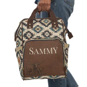 May include: A patterned backpack with a brown leather pocket featuring the name "SAMMY" in white. The pocket also displays a longhorn illustration. The bag has brown handles and accents, and a geometric pattern in shades of blue, beige, and brown.