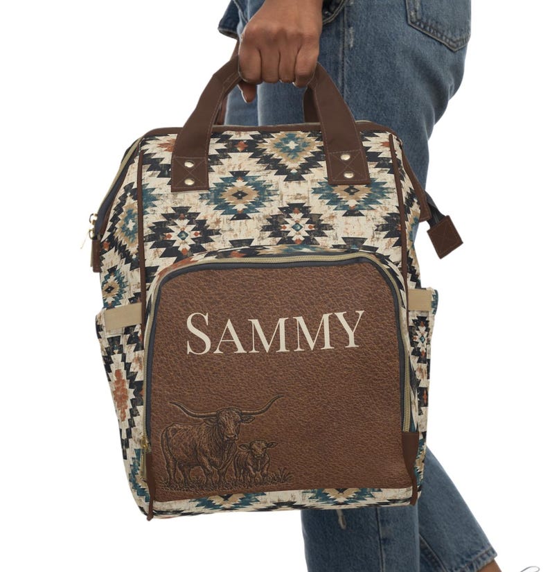 May include: A patterned backpack with a brown leather pocket featuring the name "SAMMY" in white. The pocket also displays a longhorn illustration. The bag has brown handles and accents, and a geometric pattern in shades of blue, beige, and brown.
