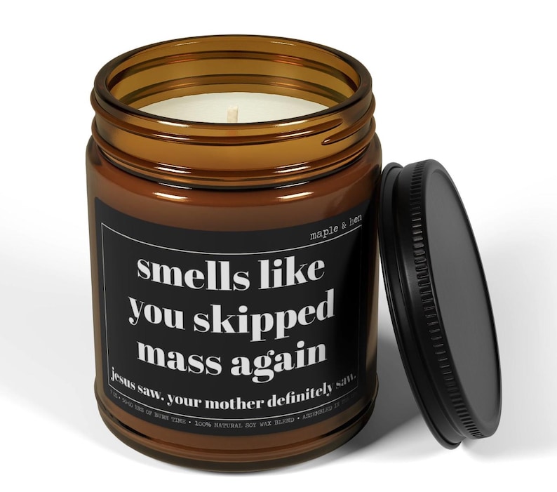 May include: A brown glass jar candle with a black lid, featuring the text "smells like you skipped mass again." The candle has a white wax interior and a label that also includes the text "Jesus saw. your mother definitely saw."