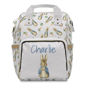 May include: White baby backpack with a repeating pattern of Peter Rabbit, carrots, and cabbage. The front pocket features the name "Charlie" in blue script above an illustration of Peter Rabbit in a blue jacket.