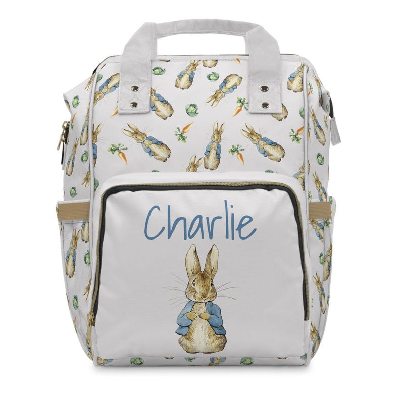 May include: White baby backpack with a repeating pattern of Peter Rabbit, carrots, and cabbage. The front pocket features the name "Charlie" in blue script above an illustration of Peter Rabbit in a blue jacket.