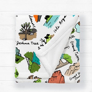 Puede incluir: Manta blanca con un dise&ntilde;o de mapa ilustrado colorido con puntos de referencia como Joshua Tree y Grand Teton. La manta tiene una textura suave y afelpada y est&aacute; doblada.